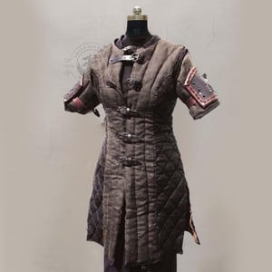 May include: A brown quilted medieval-style tunic with short sleeves, displayed on a mannequin. The tunic features leather straps and buckles, with additional leather accents on the shoulders. The garment has a diamond pattern and a split front.