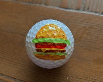 May include: A white golf ball decorated with a colourful burger design. The burger has a golden bun, green lettuce, red tomato, and a brown patty. The ball is on a wooden surface.