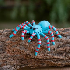 May include: A whimsical, light blue spider figurine with red accents on its legs and a pair of large, black eyes. The spider is perched on a piece of textured, brown bark, creating a playful contrast of colors and textures. This decorative item is perfect for Halloween or a quirky home decor.