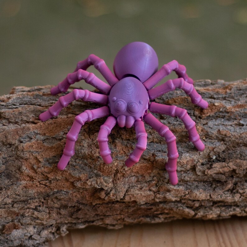 May include: A purple and pink spider figurine with eight legs, a rounded body, and detailed eyes. The spider is resting on a piece of brown bark, creating a contrast of colors and textures. The figurine is likely a decorative item or toy.