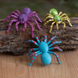 May include: Three colorful toy spiders in purple, green, and blue with red accents. The spiders are resting on a piece of wood. The spiders have multiple legs and a round body. The spiders are a fun and decorative item.