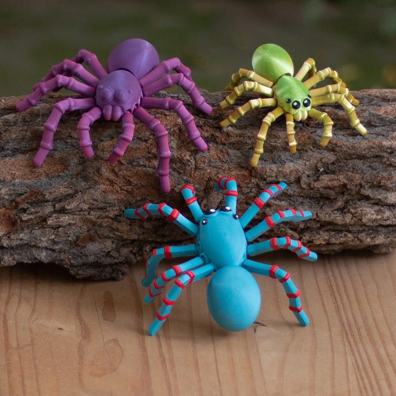 May include: Three colorful toy spiders in purple, green, and blue with red accents. The spiders are resting on a piece of wood. The spiders have multiple legs and a round body. The spiders are a fun and decorative item.