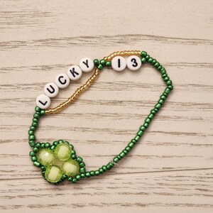 May include: A beaded bracelet with the words "LUCKY 13". The bracelet features green beads, a gold-colored beaded section, and a four-leaf clover design with light green beads. The letters and numbers are white.