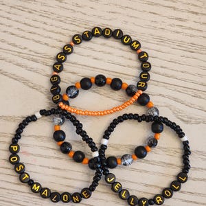 Puede incluir: Tres pulseras de cuentas con cuentas negras y detalles en naranja. Una pulsera deletrea "BIG ASS STADIUM TOUR" en cuentas de letras amarillas. Las otras dos pulseras deletrean "POST MALONE" y "JELLY ROLL".