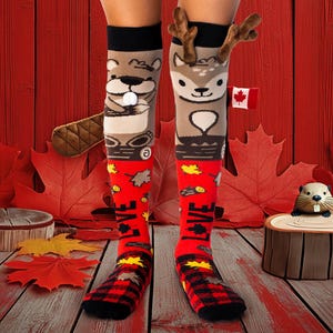 Pode incluir: Meias de cano alto com tema canadense. Uma meia apresenta um desenho de castor, a outra um veado, com uma pequena bandeira canadense. A parte inferior das meias é vermelha com folhas de bordo e a palavra "LOVE". As meias têm um padrão xadrez preto e vermelho no pé.