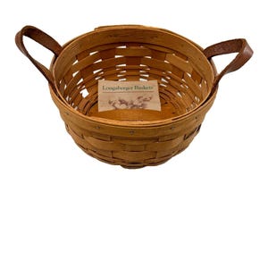 Puede incluir: Una cesta redonda de madera tejida con un acabado marrón claro y dos asas de cuero marrón unidas a los lados. Dentro de la cesta, se ve una etiqueta de papel rectangular con el texto Longaberger Baskets impreso en tinta verde oscuro.