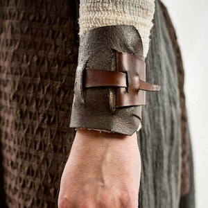 May include: A dark brown leather wrist cuff with a buckle closure. The cuff is worn over a light-colored fabric sleeve. The background features textured brown and gray fabrics.