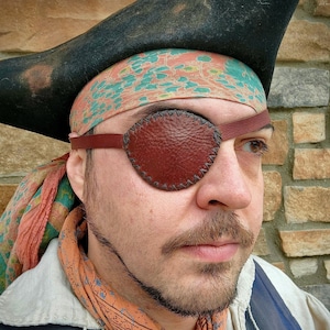 May include: A person wearing a black pirate hat, a patterned bandana, and a brown leather eye patch.