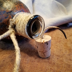 May include: A brown glass bottle with a cork stopper. The bottle is wrapped with twine and has a black rim around the opening.