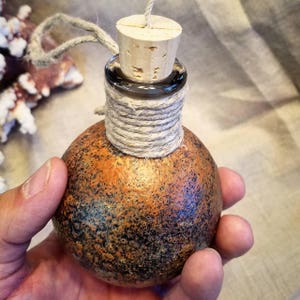 May include: A small, round, glass bottle with a cork stopper. The bottle is decorated with a rustic, textured, orange and black paint finish. The bottle is tied with a natural twine.