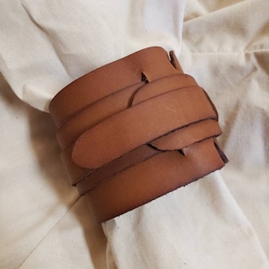 May include: A brown leather bracelet with a woven design. The bracelet is made of multiple strips of leather that are interwoven to create a unique and stylish look.