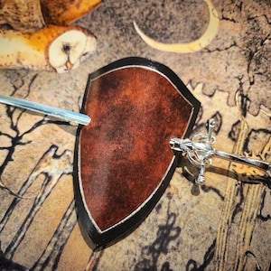 May include: A brown shield with a black border is pierced by a silver sword and a silver dagger. The shield has a textured surface. The background features an owl illustration.