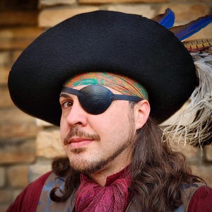May include: A man wearing a black tricorn hat with a feather, a patterned bandana, and a black leather eye patch. He has long brown hair and a beard.