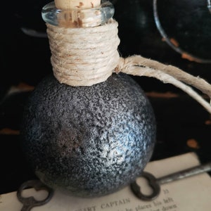 May include: A small, round, gray bottle with a cork stopper and a twine closure. The bottle is sitting on a piece of paper with text that reads "THE BRAGGART CAPTAIN".