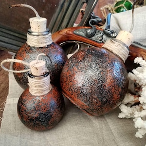 May include: Three small, round, black and orange glass bottles with cork stoppers and twine wrapped around the necks. The bottles are sitting on a light brown fabric surface.