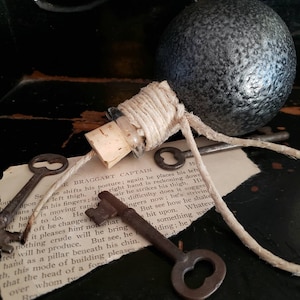 May include: A small glass bottle with a cork stopper and a twine closure. The bottle is tied to a set of old keys with a twine string. The keys are on a piece of paper with text that reads "The Braggart Captain".