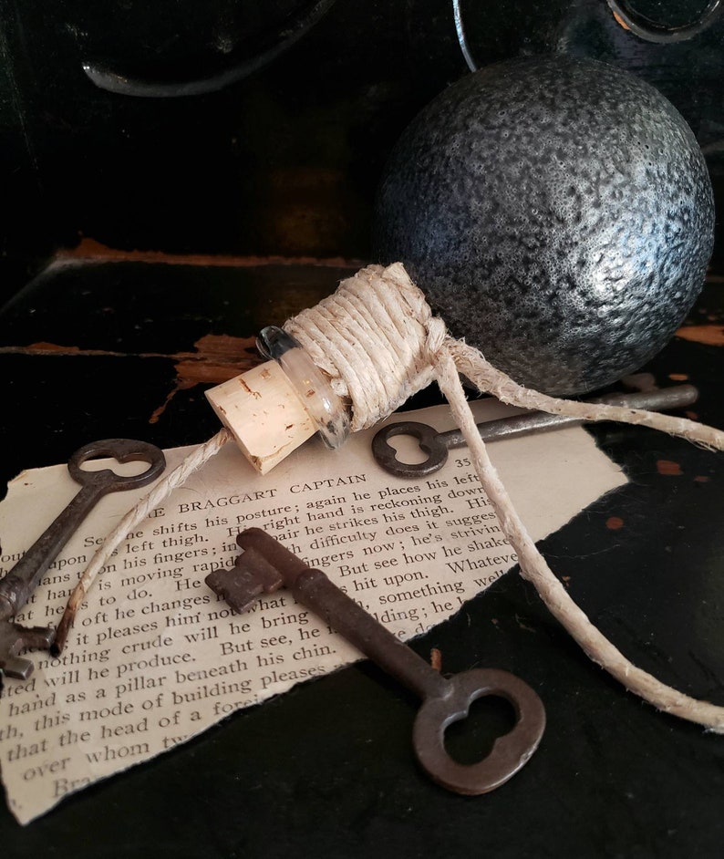 May include: A small glass bottle with a cork stopper and a twine closure. The bottle is tied to a set of old keys with a twine string. The keys are on a piece of paper with text that reads "The Braggart Captain".