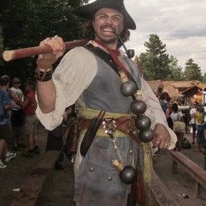 May include: A man dressed as a pirate with a black tricorn hat, a white shirt, a grey vest, and a brown belt with a gold buckle. He is holding a wooden staff in his right hand and has a pipe in his mouth. He is wearing a necklace of small round objects.