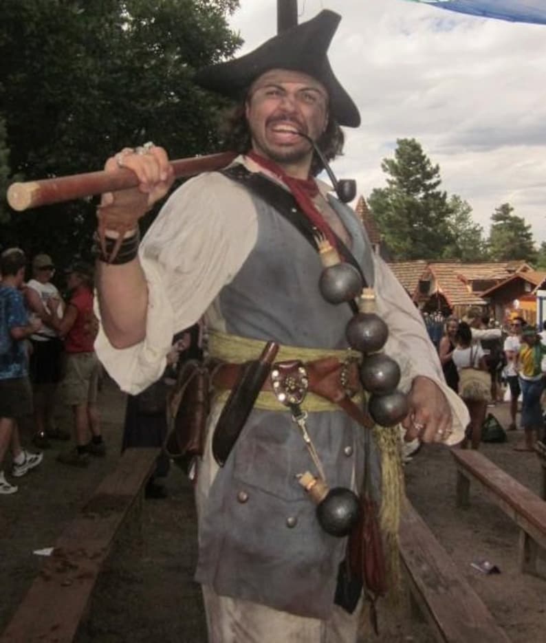 May include: A man dressed as a pirate with a black tricorn hat, a white shirt, a grey vest, and a brown belt with a gold buckle. He is holding a wooden staff in his right hand and has a pipe in his mouth. He is wearing a necklace of small round objects.
