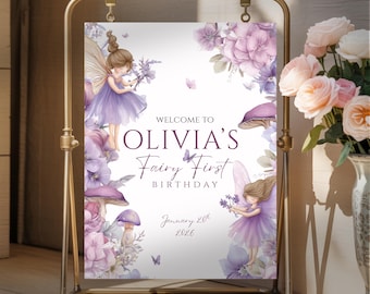 May include: A sign for a first birthday party, featuring a fairy theme. The sign has a gold metal frame and displays the text "Welcome to Olivia's Fairy First Birthday" with the date January 20th, 2026. The design includes illustrations of fairies, flowers, and mushrooms in shades of purple.