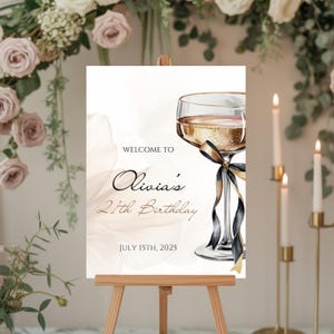 May include: A sign on an easel with the text "Welcome to Olivia's 21th Birthday July 15th, 2025". The sign features a champagne glass with black and gold ribbon. The background has a floral design and candles.