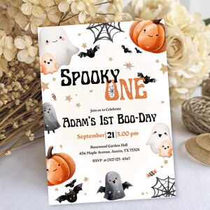 May include: A Halloween party invitation with the words "Spooky One" in black and orange. The invitation features illustrations of ghosts, bats, pumpkins, and spiderwebs. The text reads "Adam's 1st Boo-Day" and includes event details.