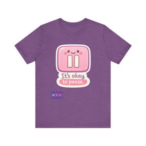 May include: Purple t-shirt featuring a pink pause button graphic with a smiling face and the text "It's okay to pause." Below the text is the phrase "THE FLOW COMPLEX" with three blue hearts.