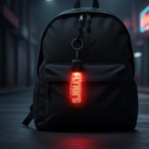 以下が含まれることがあります： フロントポケットとキーホルダーが付いた黒いバックパック。キーホルダーには、白い文字で「FLYNN'S ARCADE」と書かれた赤い光る長方形のタグが付いています。背景は暗く、ぼやけています。