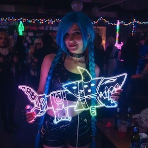 May include: A neon light sculpture in the shape of a stylized shark, with blue, pink, and yellow illumination. The sculpture is held by a person with blue hair and a black outfit. The background is a dimly lit bar with string lights.