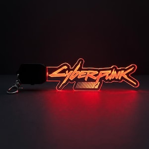 Puede incluir: Un llavero de acrílico rojo brillante con la palabra Cyberpunk en una fuente estilizada. El diseño incluye una base rectangular negra con un cierre de mosquetón de metal plateado, emitiendo una luz roja brillante que ilumina el texto.