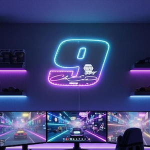May include: A neon sign in the shape of the number 9 with the number 0 inside. The sign is outlined in blue and pink, with the words "NAPA Racing" and a signature below. The sign is mounted on a purple wall above computer monitors.