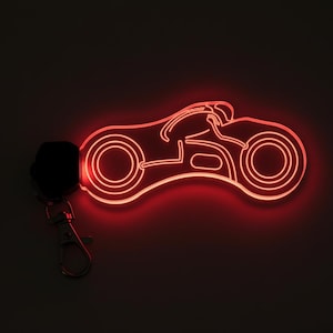 Puede incluir: Un llavero de motocicleta de neón rojo con un llavero negro y un cierre plateado. El diseño de la motocicleta presenta un piloto y dos ruedas, iluminadas con un brillo rojo brillante sobre un fondo oscuro.