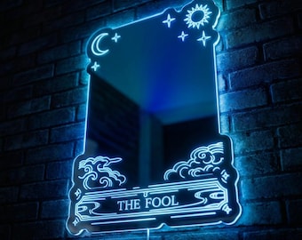 May include: A rectangular mirror with a neon blue outline, featuring celestial symbols and the text "THE FOOL." The mirror is mounted on a dark brick wall. The design includes a crescent moon, sun, stars, and clouds.
