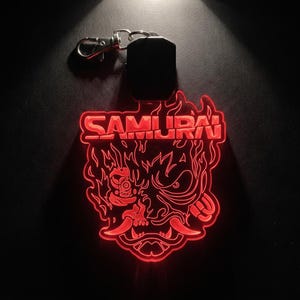 Könnte beinhalten: Ein leuchtend roter Acryl-Schlüsselanhänger mit dem Wort SAMURAI in fetter Schrift über einer stilisierten, aggressiven Maskengrafik mit spitzen Zähnen und flammenartigen Details, inklusive eines silberfarbenen Metallkarabiners.