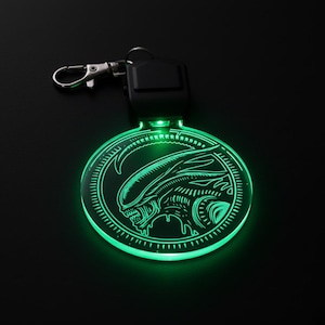 Pode incluir: Um chaveiro circular verde brilhante com um design de cabeça alienígena. O chaveiro tem um fecho prateado e uma unidade de controle preta. A ilustração alienígena é detalhada e está dentro de uma moldura circular.