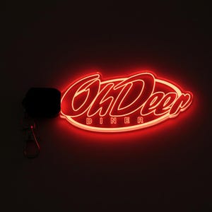 May include: A red neon sign with the words "Oh Deer Diner" in a retro font. The sign is attached to a black keychain with a silver clasp. The sign glows brightly against a dark background.