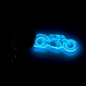 Könnte beinhalten: Ein leuchtend blaues Motorrad-Schlüsselanhänger vor schwarzem Hintergrund. Das Motorraddesign wird von einem hellblauen Neonlicht beleuchtet, das die Konturen des Fahrzeugs hervorhebt. Der Schlüsselanhänger ist an einem dunklen, unbeleuchteten Objekt befestigt.