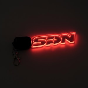 May include: A glowing red acrylic keychain with the letters "SDN". The keychain is illuminated with a bright red light, and is attached to a black fob and a silver clasp. The letters are in a bold, modern font.