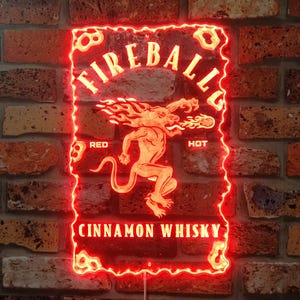 Op de afbeelding: Rood neonbord met de tekst "Fireball" en "Cinnamon Whisky". In het midden staat een vurige creatuur. Het bord is bevestigd aan een bakstenen muur. De rand van het bord heeft een decoratieve, golvende rand.