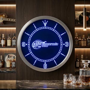 May include: A round, illuminated wall clock with a silver frame. The clock face is a deep blue with the words "Miller Time LIVE!" in white. The clock has white hour markers and a silver outer ring. The background is a dark wood.