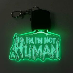 Könnte beinhalten: Ein grün leuchtender Schlüsselanhänger mit dem Text "NO, I'M NOT A HUMAN". Der Schlüsselanhänger hat einen silbernen Verschluss und eine schwarze Steuerbox. Der Text ist in einer verwitterten Schriftart.