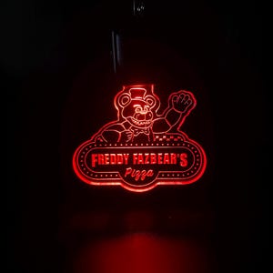 Könnte beinhalten: Ein rotes Neon-Schild mit dem Logo von Freddy Fazbear's Pizza. Das Schild zeigt einen winkenden Bärencharakter mit dem Text "Freddy Fazbear's Pizza" in einer stilisierten Schriftart. Das Schild ist vor einem dunklen Hintergrund beleuchtet.