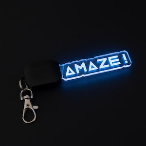 Pode incluir: Um porta-chaves preto com um pendente retangular azul brilhante. O pendente tem a palavra "AMAZE!" em letras maiúsculas brancas. O porta-chaves inclui um fecho preto e um porta-chaves prateado.
