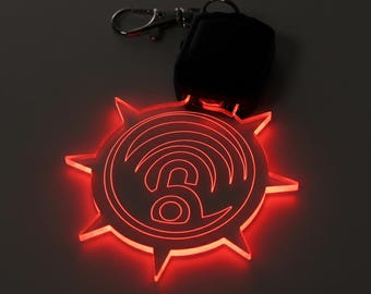 May include: A red, illuminated keychain with a unique design. The keychain features a circular shape with pointed edges and a central design resembling a stylized '6' with radiating lines. A black remote and silver keyring are attached.