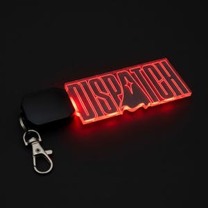 May include: A rectangular keychain with the word "DISPATCH" in red neon lights. The keychain has a black backing and a silver clasp. The background is black.