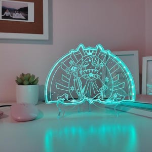 May include: A clear acrylic LED lamp featuring a cartoon character design with a crown and scepter. The lamp emits a bright teal glow and is supported by a clear stand. The design includes stars and decorative elements, creating a whimsical aesthetic.