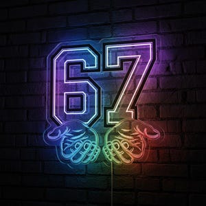 Puede incluir: Un letrero de neón con el número "67" en una fuente universitaria, con manos ahuecadas debajo. El letrero brilla con un degradado de colores, incluyendo azul, morado y rojo, contra una pared de ladrillos oscuros.