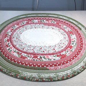 May include: An oval-shaped fabric placemat constructed from coiled strips of cotton material. The design features concentric rings of floral patterns in shades of red, white, and green, with a central white section decorated with small red dots.
