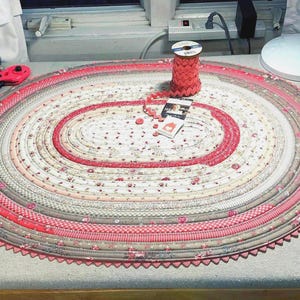 May include: An oval-shaped coiled fabric mat featuring concentric rings of floral, gingham, and solid pink and grey patterns. A spool of pink rickrack trim, a small tag, and a few matching buttons are placed on the center of the mat.