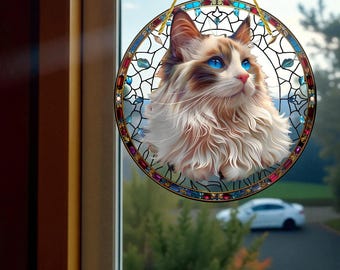 May include: A round stained glass window decoration featuring a fluffy cat with blue eyes. The cat is brown, white, and tan, set against a colorful stained glass border with a gold hanging cord.